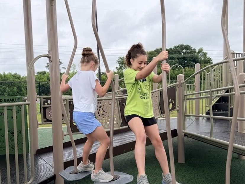 Two girls, one in a lime Motivate shirt, stepping across balance pods on an outdoor playground