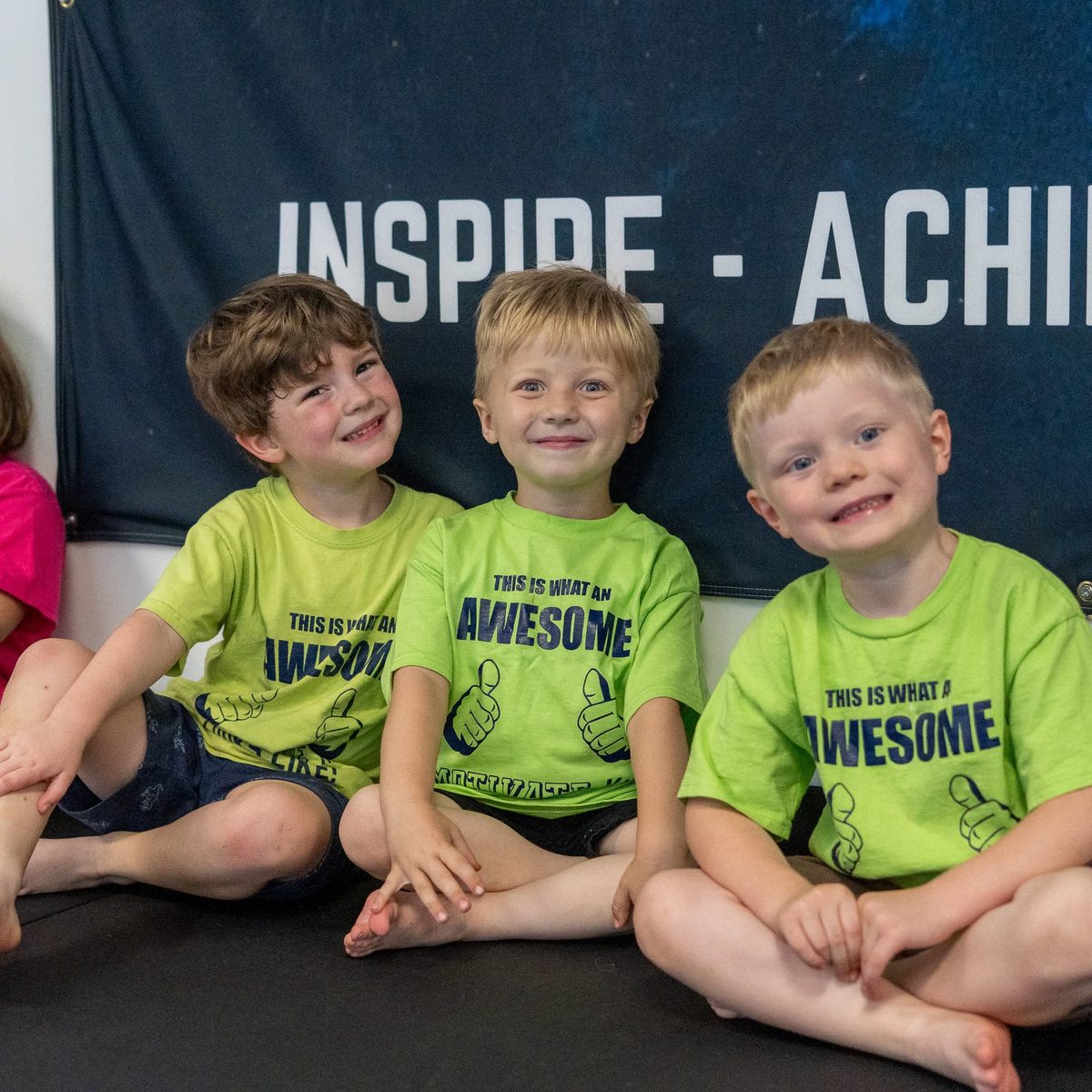 Three smiling boys in lime Motivate shirts sitting on the mat under the Inspire Achieve Succeed banner