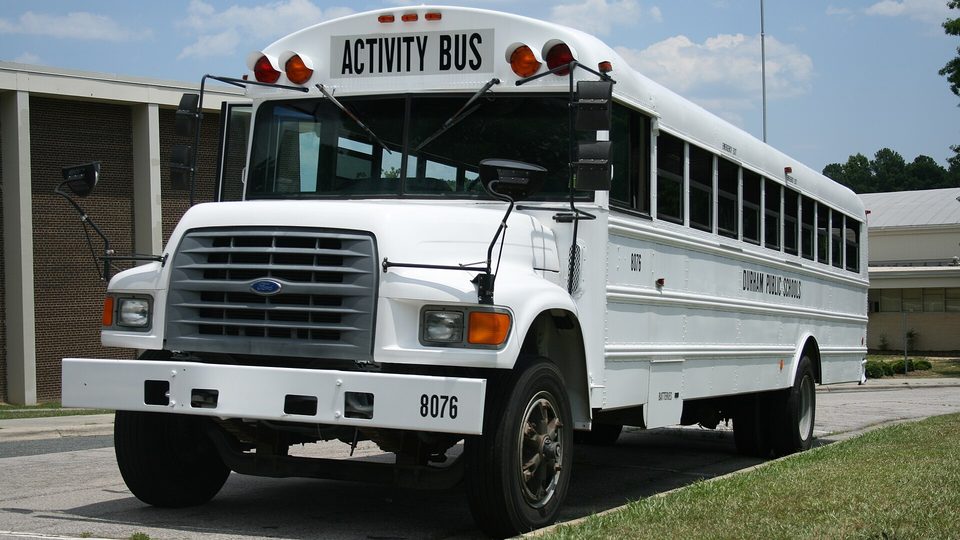 A white activity bus parked outside a brick school building on a sunny day